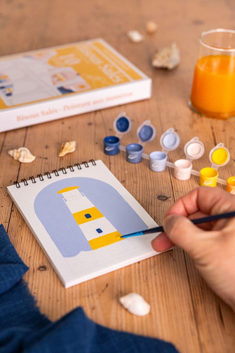 peinture par numéros d'un phare en cours de réalisation avec pots de peinture et verre de jus d'orange sur table en bois